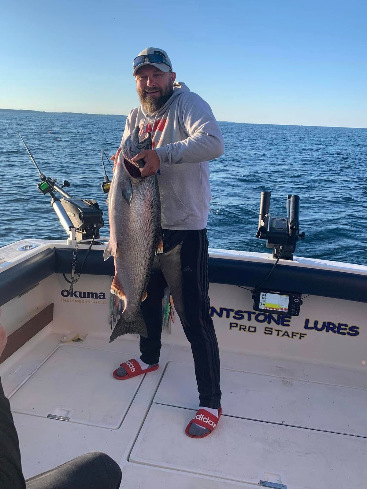 A bearded fisherman holding a large lake trout aboard a charter boat rigged with Okuma gear
