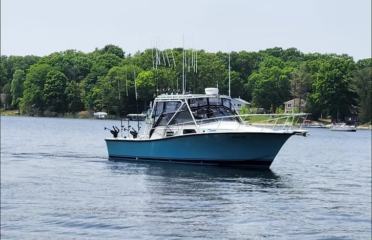 A teal hulled charter boat lying quiet on green water with the rods racked and a wooded shore behind