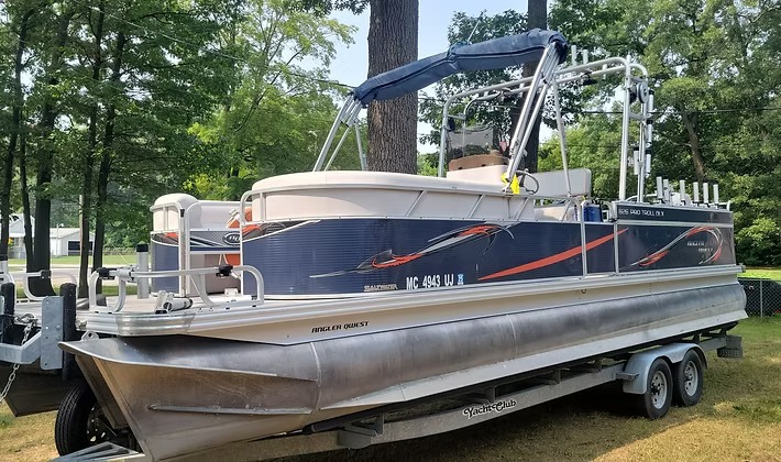 A fishing pontoon rigged with rod holders and a trolling spread, parked on its trailer under the trees